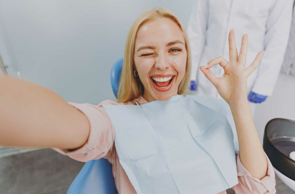 A smiling young woman in a dental chair winking and taking a selfie with her right hand, while holding up her left hand giving an okay symbol.