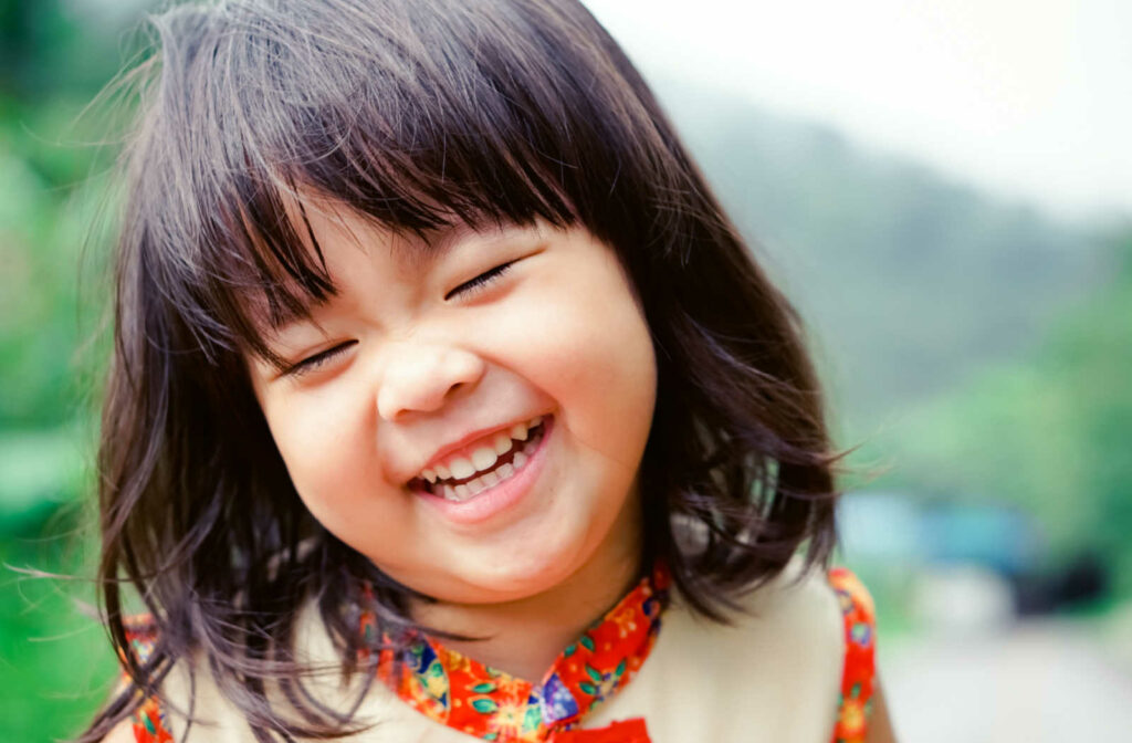 A portrait of a young Asian girl smiling and laughing outside with her eyes closed.