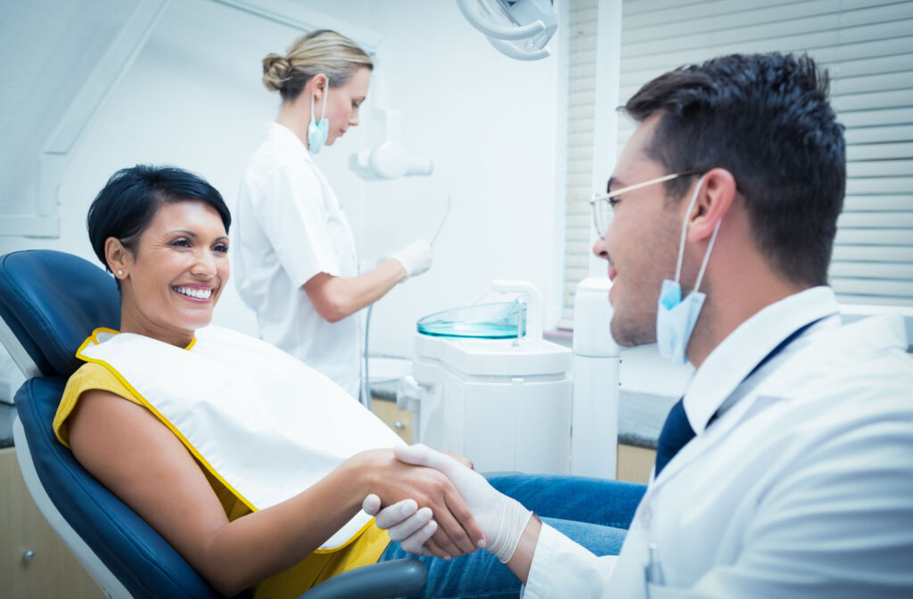 A woman in a dentist's office smiling and shaking hands with her dentist while a dental assistant is working in the background.