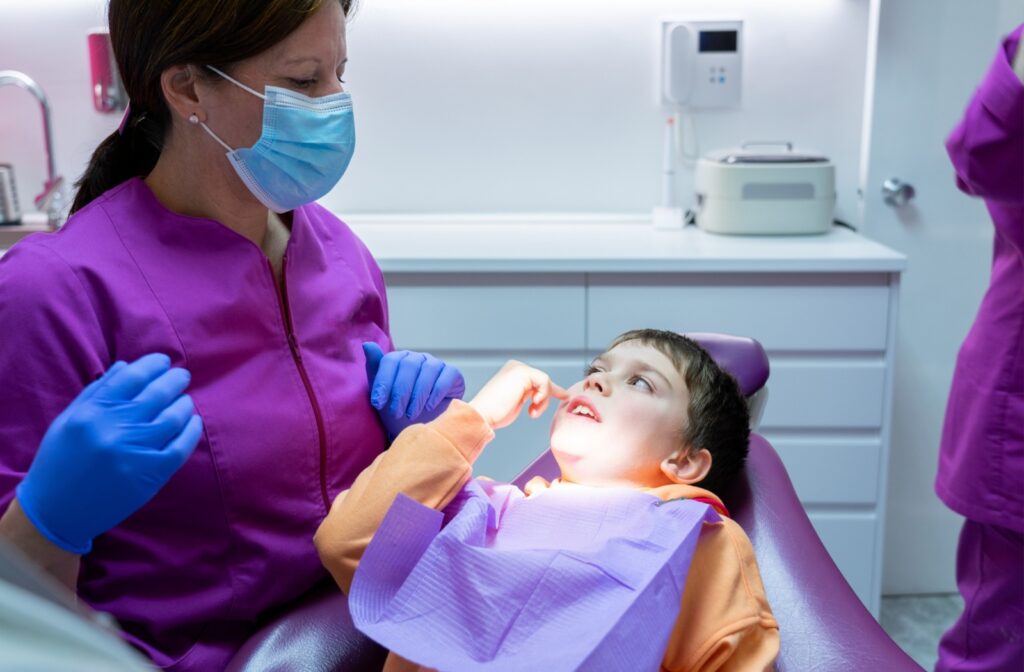 A child sits in a dentist's chair pointing to his teeth while a dentist listens